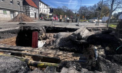 Großes Loch in einer Straße mit freigelegten Rohren und abgesacktem Asphalt; die Baustelle ist abgesperrt, im Hintergrund stehen Bagger und Gebäude.
