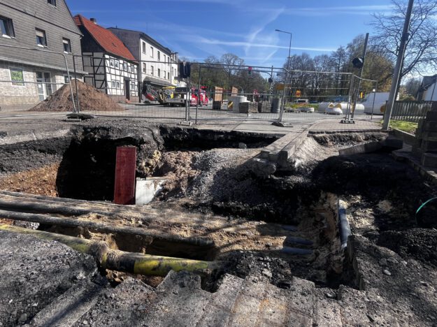 Großes Loch in einer Straße mit freigelegten Rohren und abgesacktem Asphalt; die Baustelle ist abgesperrt, im Hintergrund stehen Bagger und Gebäude.