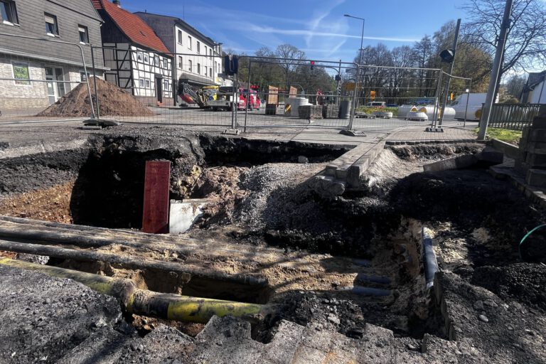 Großes Loch in einer Straße mit freigelegten Rohren und abgesacktem Asphalt; die Baustelle ist abgesperrt, im Hintergrund stehen Bagger und Gebäude.