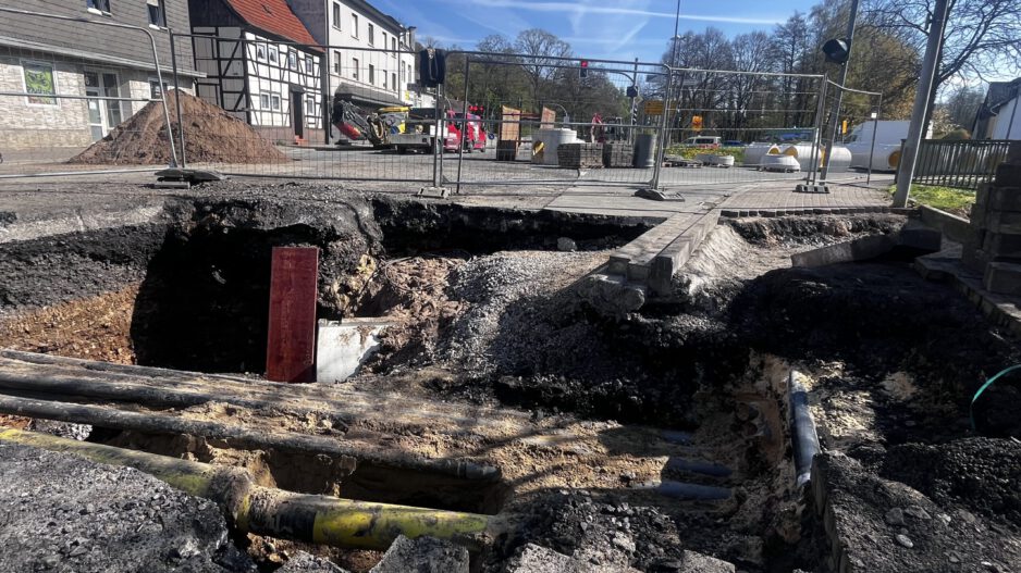 Großes Loch in einer Straße mit freigelegten Rohren und abgesacktem Asphalt; die Baustelle ist abgesperrt, im Hintergrund stehen Bagger und Gebäude.