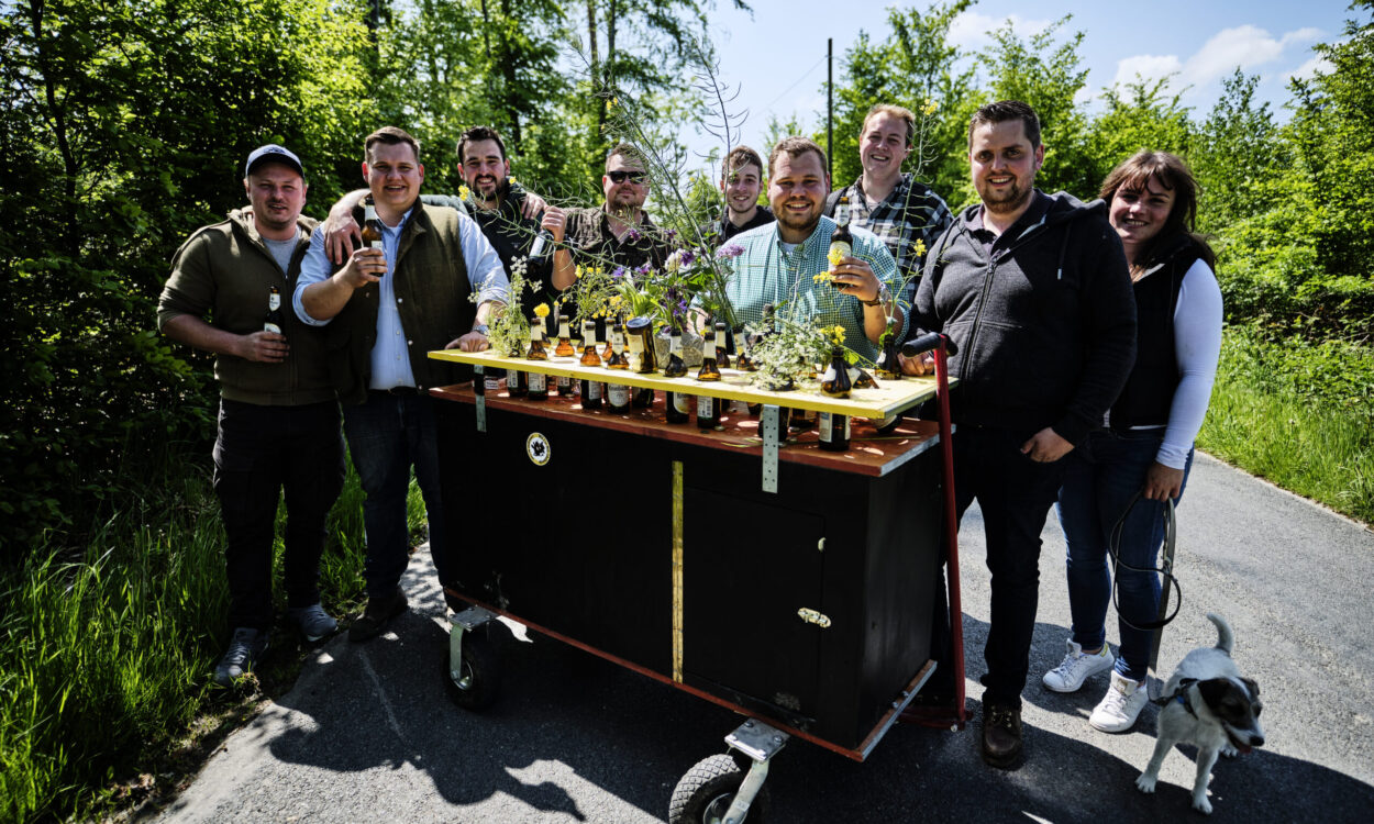Eine Gruppe junger Erwachsener steht auf einem Waldweg um einen geschmückten Bollerwagen mit Getränken und Blumen; einige halten Flaschen in der Hand, daneben läuft ein kleiner Hund.