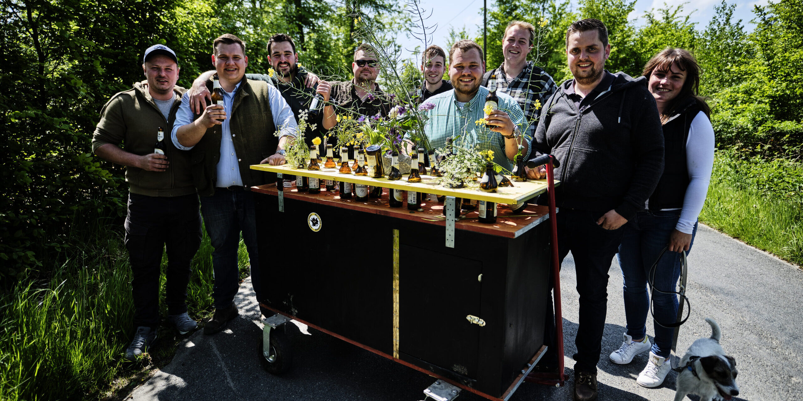 Eine Gruppe junger Erwachsener steht auf einem Waldweg um einen geschmückten Bollerwagen mit Getränken und Blumen; einige halten Flaschen in der Hand, daneben läuft ein kleiner Hund.