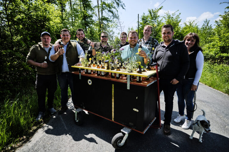 Eine Gruppe junger Erwachsener steht auf einem Waldweg um einen geschmückten Bollerwagen mit Getränken und Blumen; einige halten Flaschen in der Hand, daneben läuft ein kleiner Hund.