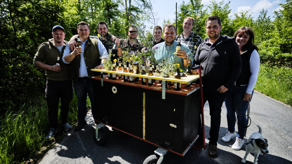 Eine Gruppe junger Erwachsener steht auf einem Waldweg um einen geschmückten Bollerwagen mit Getränken und Blumen; einige halten Flaschen in der Hand, daneben läuft ein kleiner Hund.
