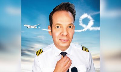 Ein Mann in Pilotenuniform schaut frontal in die Kamera und richtet seine Krawatte, während im Hintergrund ein Flugzeug am Himmel eine Schleife aus Kondensstreifen zieht.