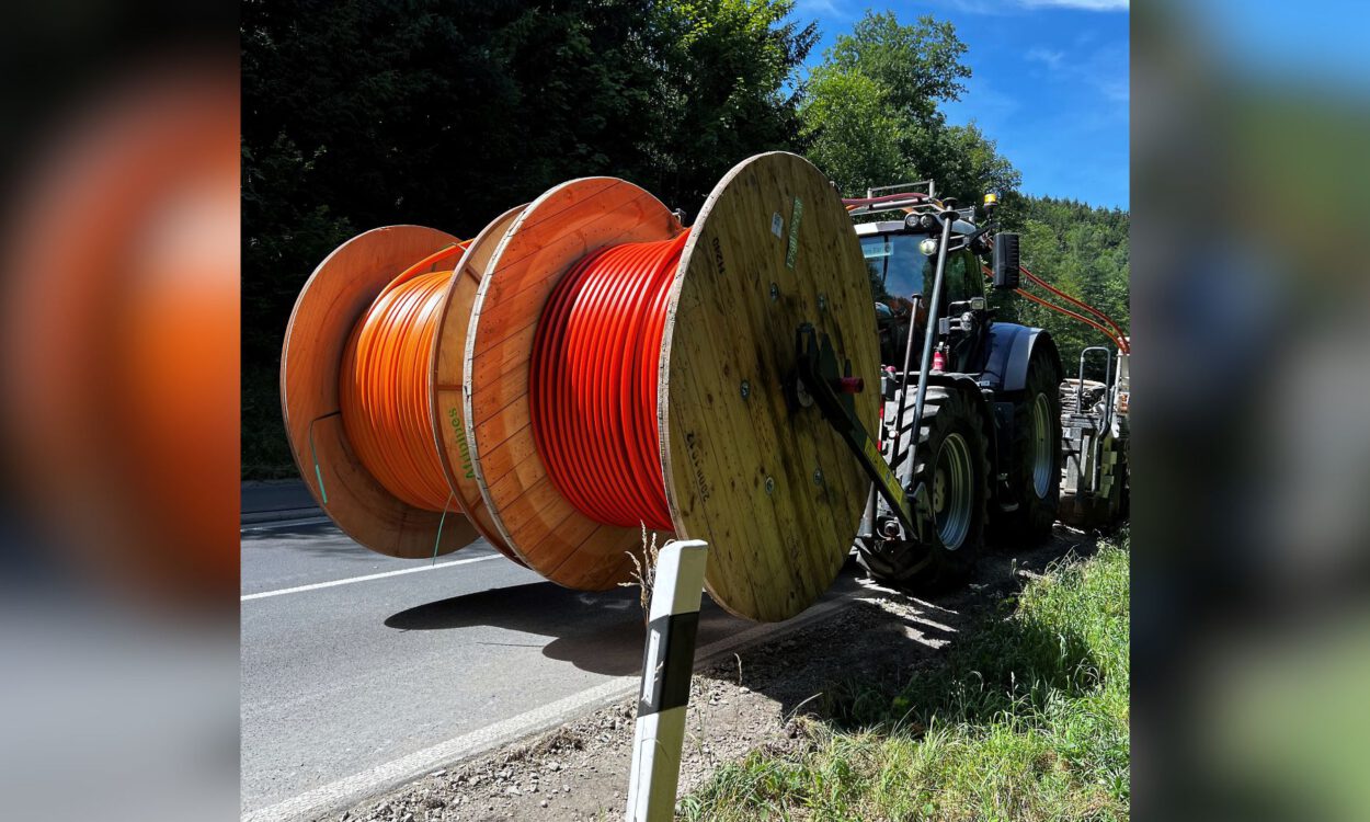 Traktor transportiert große Kabeltrommeln mit orangefarbenen Leitungen am Straßenrand entlang.