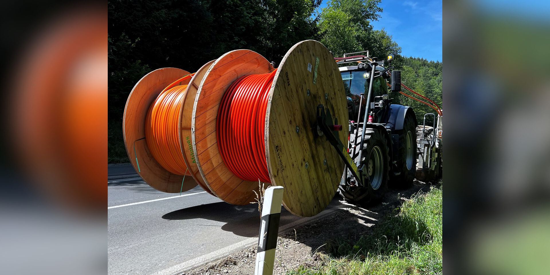 Traktor transportiert große Kabeltrommeln mit orangefarbenen Leitungen am Straßenrand entlang.