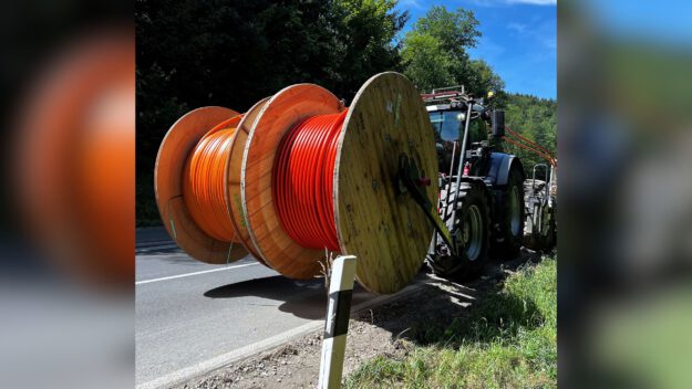 Traktor transportiert große Kabeltrommeln mit orangefarbenen Leitungen am Straßenrand entlang.