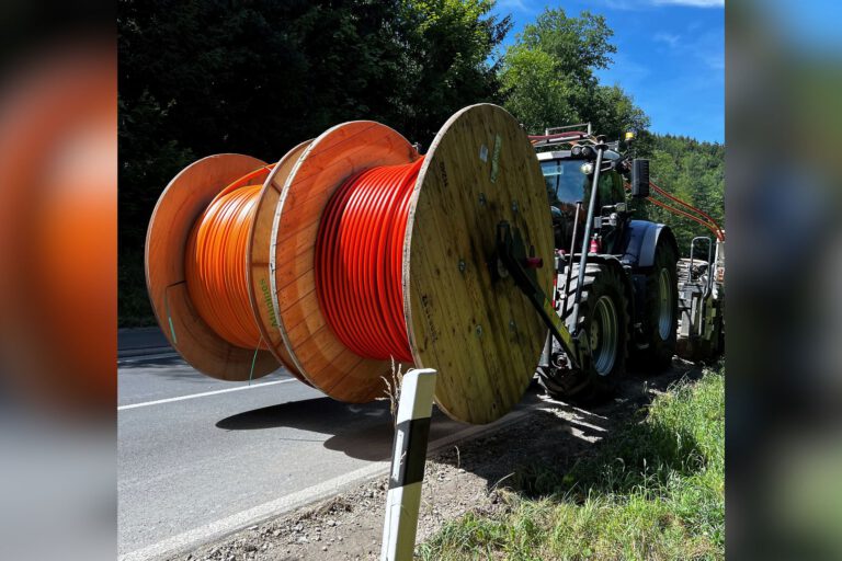 Traktor transportiert große Kabeltrommeln mit orangefarbenen Leitungen am Straßenrand entlang.