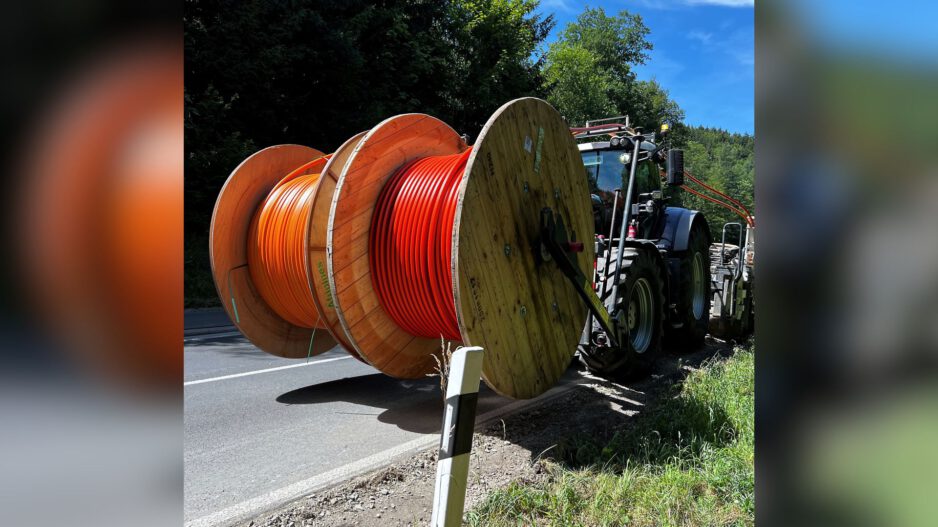 Traktor transportiert große Kabeltrommeln mit orangefarbenen Leitungen am Straßenrand entlang.