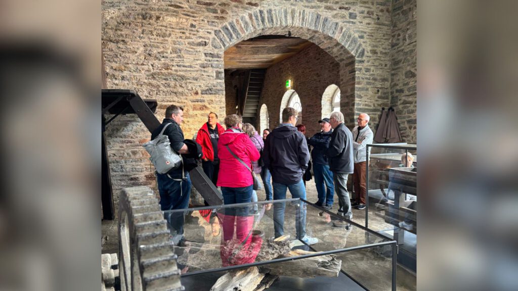 Gruppe von Menschen steht in einem historischen Gebäude mit Steinwänden und hört einer Führung zu; im Vordergrund sind Ausstellungsstücke hinter Glas zu sehen.