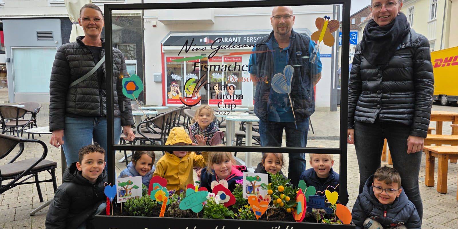 Gruppe von Kindern und Erwachsenen steht auf einem Platz hinter einem dekorierten Blumenbeet und einer Glasscheibe mit Werbeaufschrift für handwerklich hergestelltes Eis.