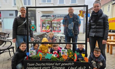 Gruppe von Kindern und Erwachsenen steht auf einem Platz hinter einem dekorierten Blumenbeet und einer Glasscheibe mit Werbeaufschrift für handwerklich hergestelltes Eis.