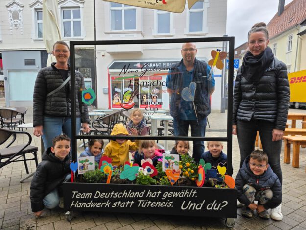 Gruppe von Kindern und Erwachsenen steht auf einem Platz hinter einem dekorierten Blumenbeet und einer Glasscheibe mit Werbeaufschrift für handwerklich hergestelltes Eis.