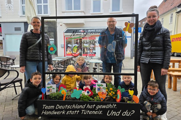 Gruppe von Kindern und Erwachsenen steht auf einem Platz hinter einem dekorierten Blumenbeet und einer Glasscheibe mit Werbeaufschrift für handwerklich hergestelltes Eis.