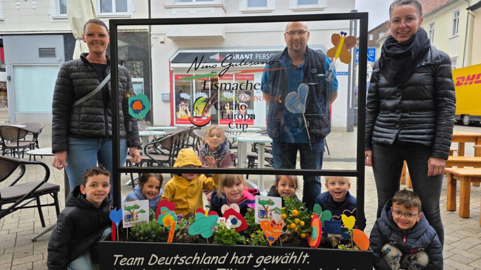 Gruppe von Kindern und Erwachsenen steht auf einem Platz hinter einem dekorierten Blumenbeet und einer Glasscheibe mit Werbeaufschrift für handwerklich hergestelltes Eis.