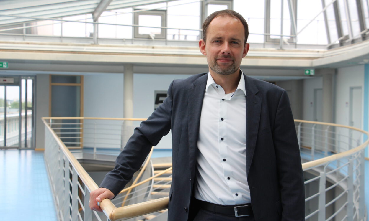 Mann im Anzug steht in einem modernen Gebäude an einem Geländer und blickt in die Kamera.