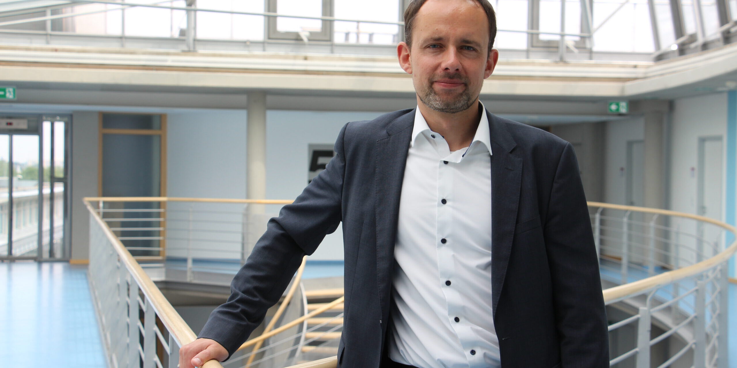 Mann im Anzug steht in einem modernen Gebäude an einem Geländer und blickt in die Kamera.