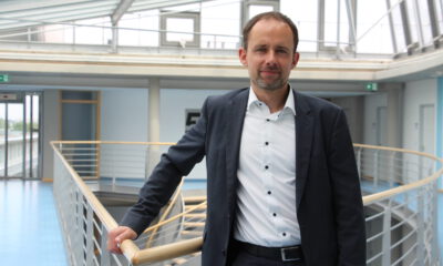 Mann im Anzug steht in einem modernen Gebäude an einem Geländer und blickt in die Kamera.