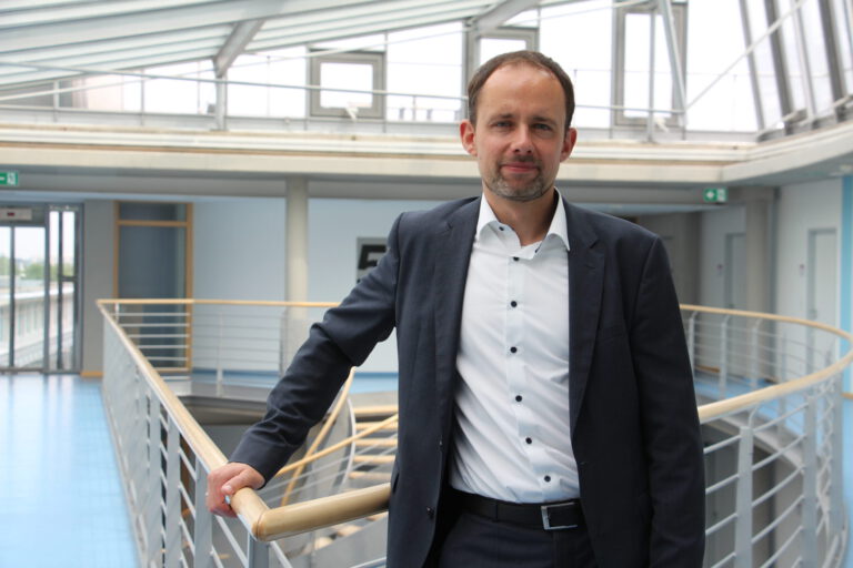 Mann im Anzug steht in einem modernen Gebäude an einem Geländer und blickt in die Kamera.