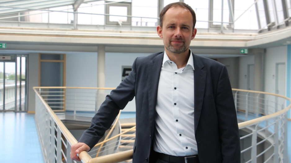 Mann im Anzug steht in einem modernen Gebäude an einem Geländer und blickt in die Kamera.