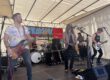 Band mit mehreren Musikern spielt live auf einer Bühne unter einem Zelt; ein Sänger mit Gitarre steht am Mikrofon, begleitet von Bassist, Schlagzeuger und weiterem Gitarristen.