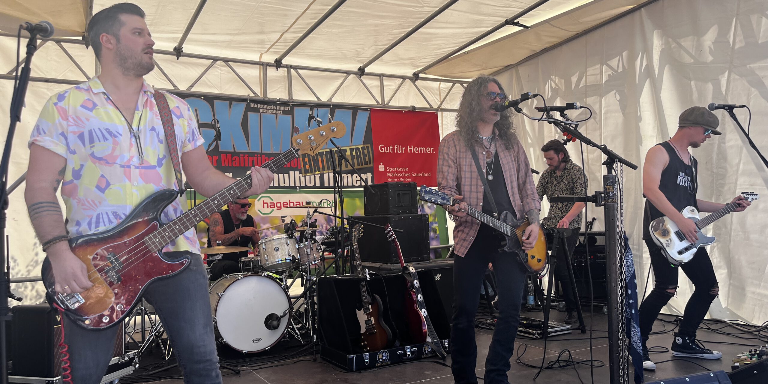 Band mit mehreren Musikern spielt live auf einer Bühne unter einem Zelt; ein Sänger mit Gitarre steht am Mikrofon, begleitet von Bassist, Schlagzeuger und weiterem Gitarristen.
