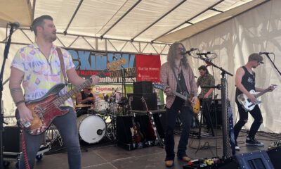 Band mit mehreren Musikern spielt live auf einer Bühne unter einem Zelt; ein Sänger mit Gitarre steht am Mikrofon, begleitet von Bassist, Schlagzeuger und weiterem Gitarristen.