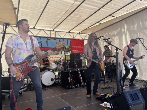 Band mit mehreren Musikern spielt live auf einer Bühne unter einem Zelt; ein Sänger mit Gitarre steht am Mikrofon, begleitet von Bassist, Schlagzeuger und weiterem Gitarristen.