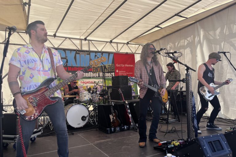 Band mit mehreren Musikern spielt live auf einer Bühne unter einem Zelt; ein Sänger mit Gitarre steht am Mikrofon, begleitet von Bassist, Schlagzeuger und weiterem Gitarristen.