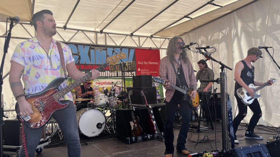 Band mit mehreren Musikern spielt live auf einer Bühne unter einem Zelt; ein Sänger mit Gitarre steht am Mikrofon, begleitet von Bassist, Schlagzeuger und weiterem Gitarristen.