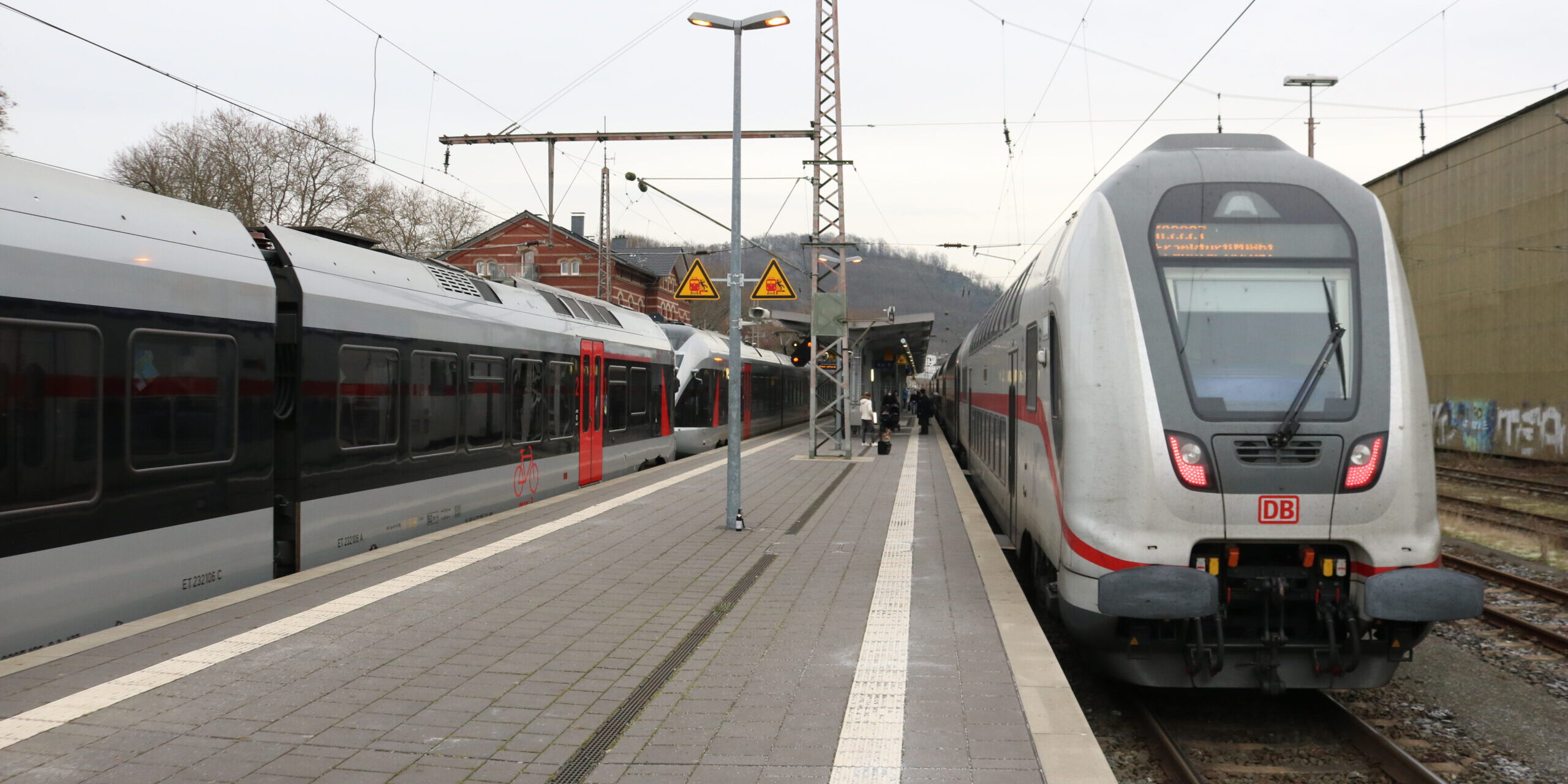 Zwei Züge der Deutschen Bahn stehen an einem Bahnsteig; Reisende gehen zwischen den Gleisen, im Hintergrund sind Bahnhofsgebäude und Oberleitungen zu sehen.