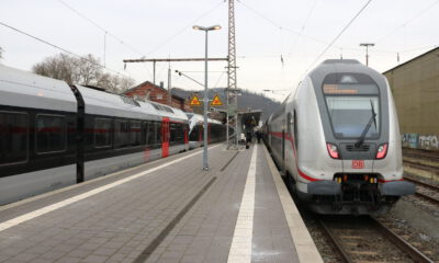 Zwei Züge der Deutschen Bahn stehen an einem Bahnsteig; Reisende gehen zwischen den Gleisen, im Hintergrund sind Bahnhofsgebäude und Oberleitungen zu sehen.