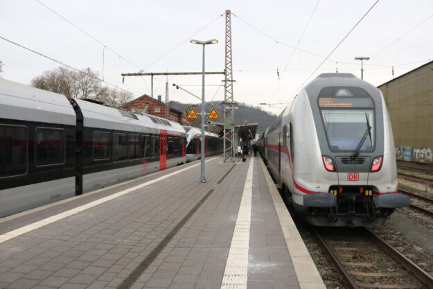 Zwei Züge der Deutschen Bahn stehen an einem Bahnsteig; Reisende gehen zwischen den Gleisen, im Hintergrund sind Bahnhofsgebäude und Oberleitungen zu sehen.