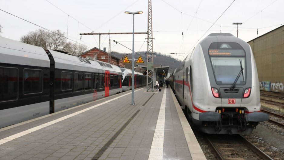 Zwei Züge der Deutschen Bahn stehen an einem Bahnsteig; Reisende gehen zwischen den Gleisen, im Hintergrund sind Bahnhofsgebäude und Oberleitungen zu sehen.