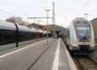 Zwei Züge der Deutschen Bahn stehen an einem Bahnsteig; Reisende gehen zwischen den Gleisen, im Hintergrund sind Bahnhofsgebäude und Oberleitungen zu sehen.
