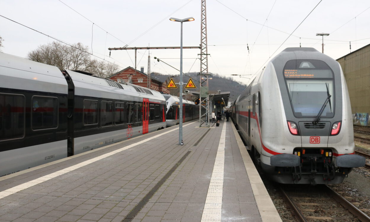 Zwei Züge der Deutschen Bahn stehen an einem Bahnsteig; Reisende gehen zwischen den Gleisen, im Hintergrund sind Bahnhofsgebäude und Oberleitungen zu sehen.