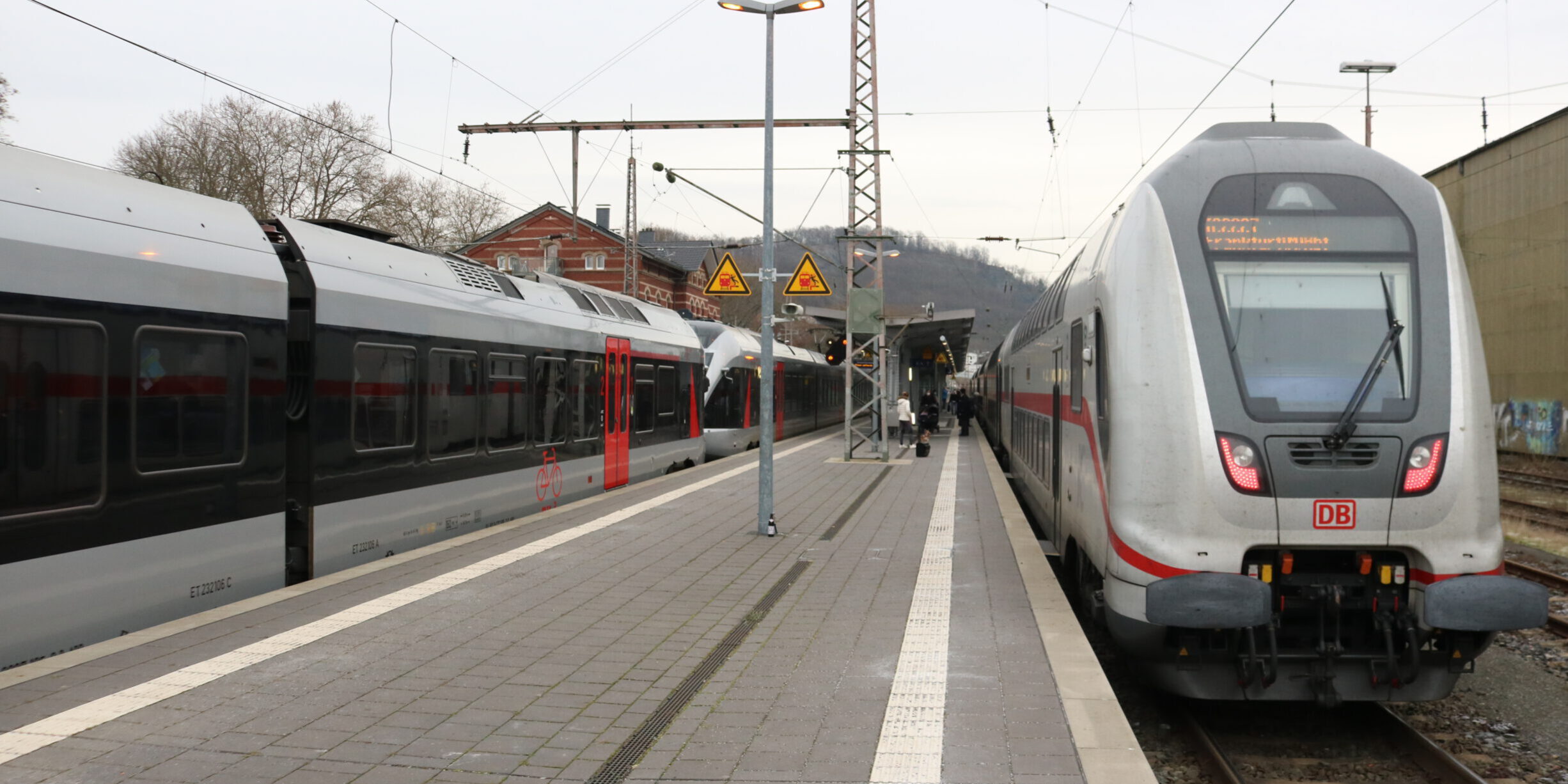 Zwei Züge der Deutschen Bahn stehen an einem Bahnsteig; Reisende gehen zwischen den Gleisen, im Hintergrund sind Bahnhofsgebäude und Oberleitungen zu sehen.