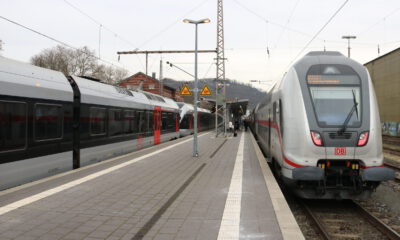 Zwei Züge der Deutschen Bahn stehen an einem Bahnsteig; Reisende gehen zwischen den Gleisen, im Hintergrund sind Bahnhofsgebäude und Oberleitungen zu sehen.