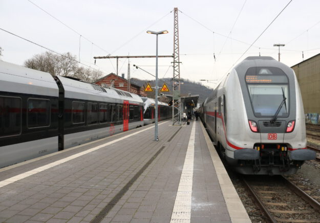Zwei Züge der Deutschen Bahn stehen an einem Bahnsteig; Reisende gehen zwischen den Gleisen, im Hintergrund sind Bahnhofsgebäude und Oberleitungen zu sehen.
