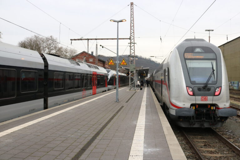 Zwei Züge der Deutschen Bahn stehen an einem Bahnsteig; Reisende gehen zwischen den Gleisen, im Hintergrund sind Bahnhofsgebäude und Oberleitungen zu sehen.