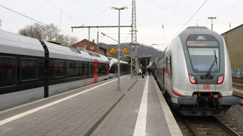 Zwei Züge der Deutschen Bahn stehen an einem Bahnsteig; Reisende gehen zwischen den Gleisen, im Hintergrund sind Bahnhofsgebäude und Oberleitungen zu sehen.