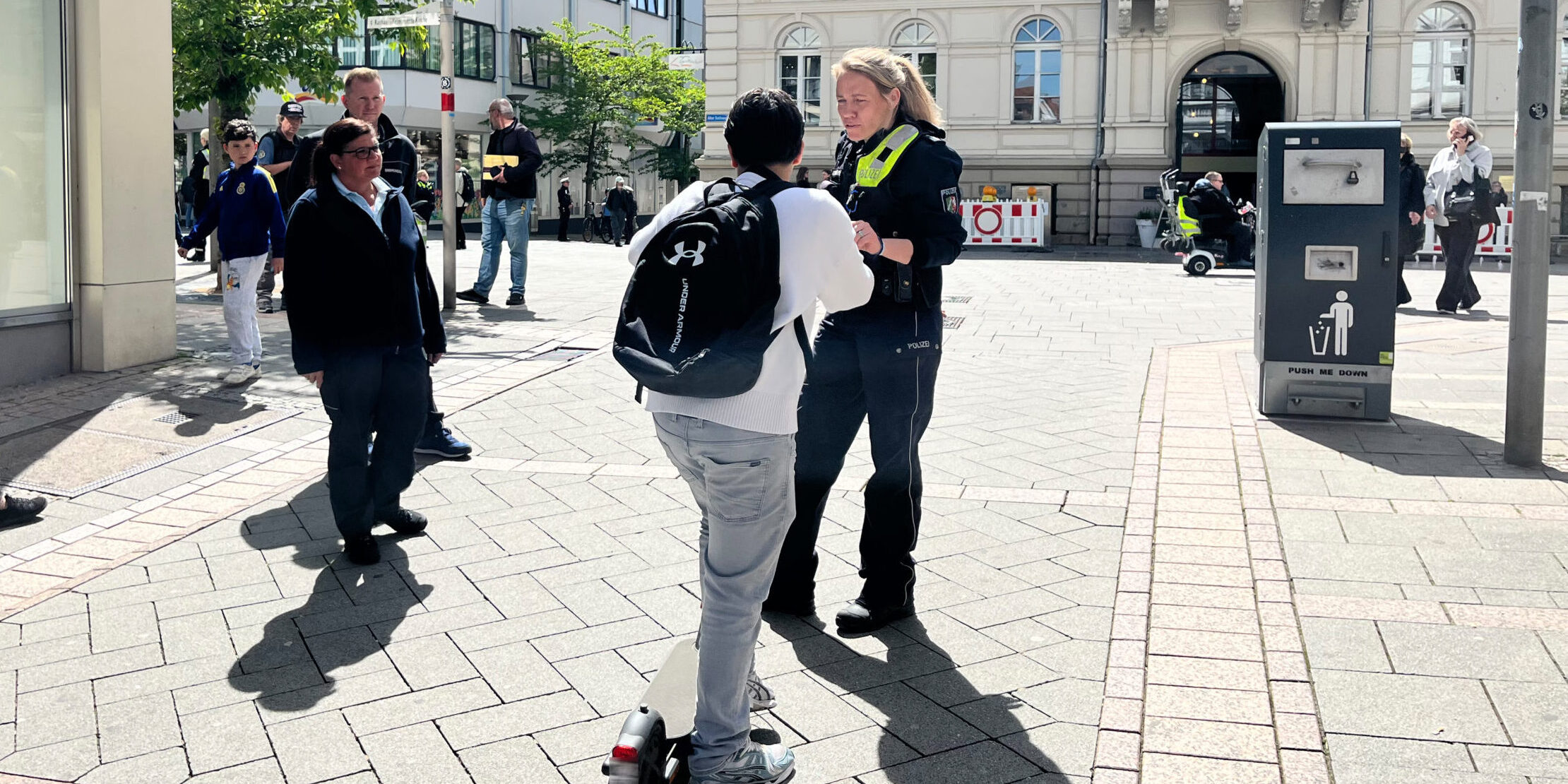 Eine Polizistin spricht auf einem sonnigen Platz vor dem Rathaus mit einer Person auf einem E-Scooter, während Passanten in der Innenstadt vorbeigehen.