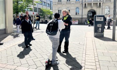 Eine Polizistin spricht auf einem sonnigen Platz vor dem Rathaus mit einer Person auf einem E-Scooter, während Passanten in der Innenstadt vorbeigehen.