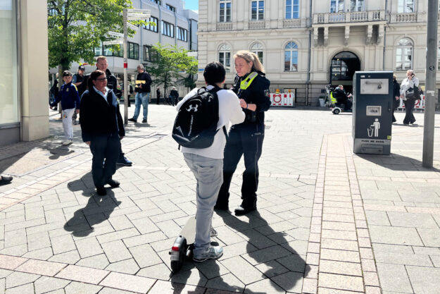 Eine Polizistin spricht auf einem sonnigen Platz vor dem Rathaus mit einer Person auf einem E-Scooter, während Passanten in der Innenstadt vorbeigehen.