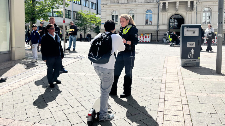 Eine Polizistin spricht auf einem sonnigen Platz vor dem Rathaus mit einer Person auf einem E-Scooter, während Passanten in der Innenstadt vorbeigehen.