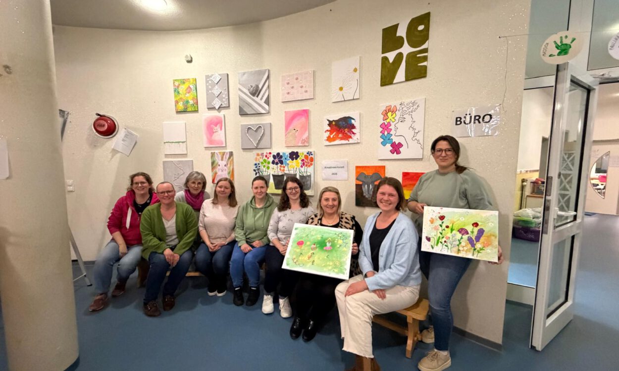 Gruppe von Frauen sitzt und steht in einem Raum vor einer Wand mit bunten Kunstwerken und hält selbst gestaltete Bilder aus einem Kreativworkshop in den Händen.