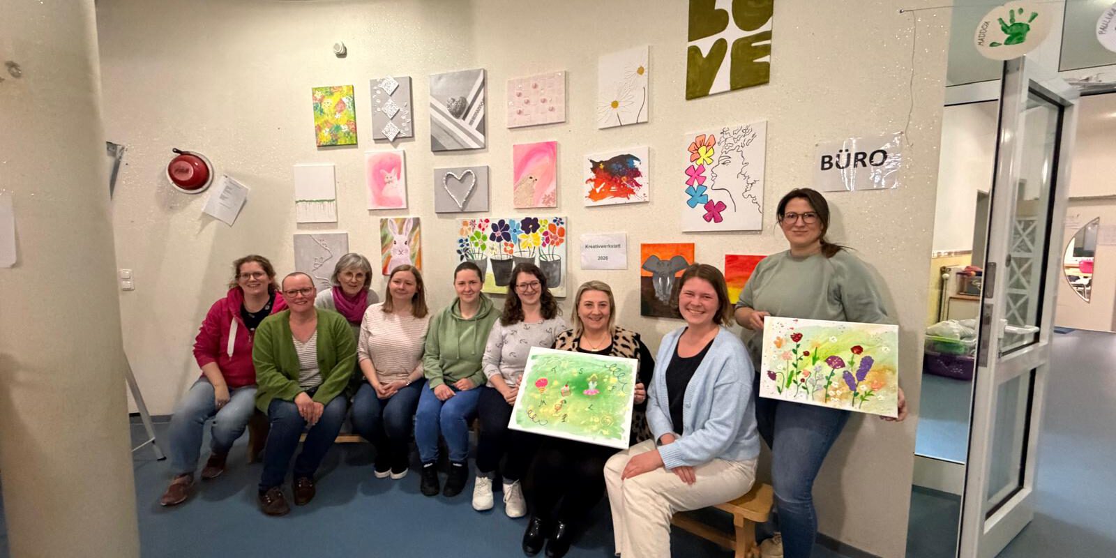 Gruppe von Frauen sitzt und steht in einem Raum vor einer Wand mit bunten Kunstwerken und hält selbst gestaltete Bilder aus einem Kreativworkshop in den Händen.