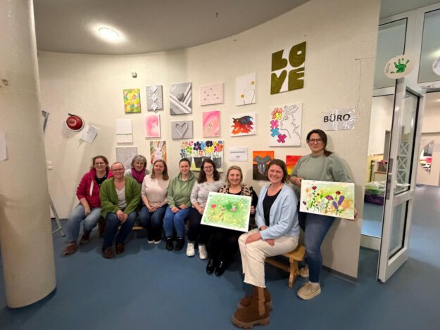 Gruppe von Frauen sitzt und steht in einem Raum vor einer Wand mit bunten Kunstwerken und hält selbst gestaltete Bilder aus einem Kreativworkshop in den Händen.