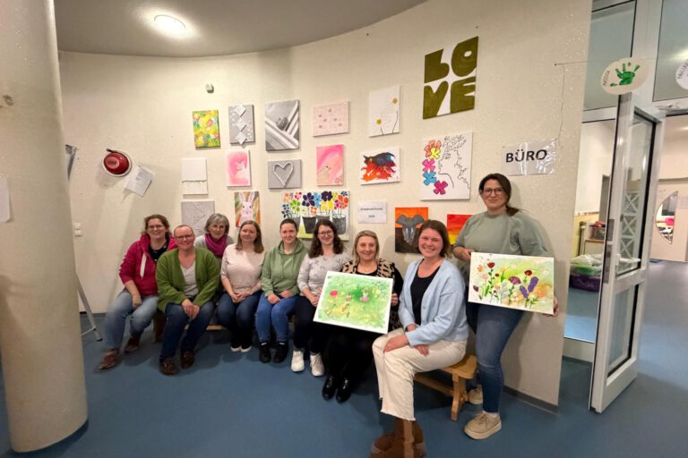 Gruppe von Frauen sitzt und steht in einem Raum vor einer Wand mit bunten Kunstwerken und hält selbst gestaltete Bilder aus einem Kreativworkshop in den Händen.